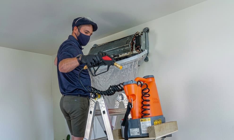 Masked technician cleans a wall-mounted air conditioning unit with a pressure sprayer and collection bag.